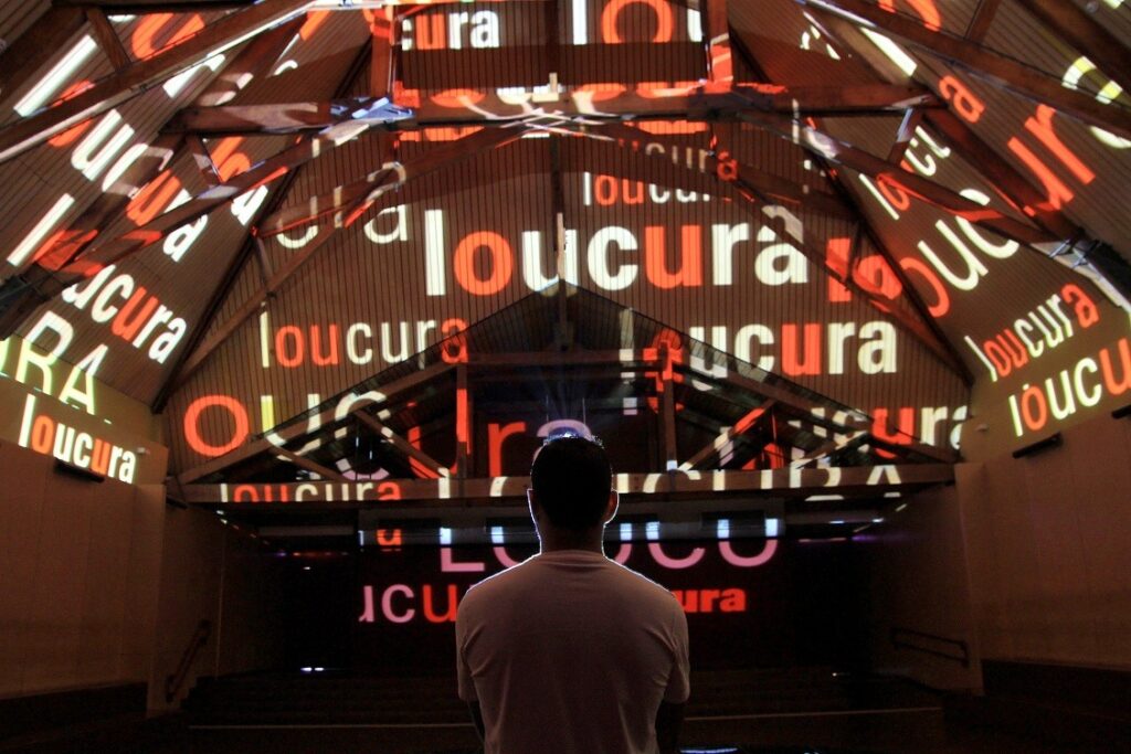 A palavra loucura projetada diversas vezes na parede, com homem olhando. Imagem ilustrativa texto Museu da Língua Portuguesa.