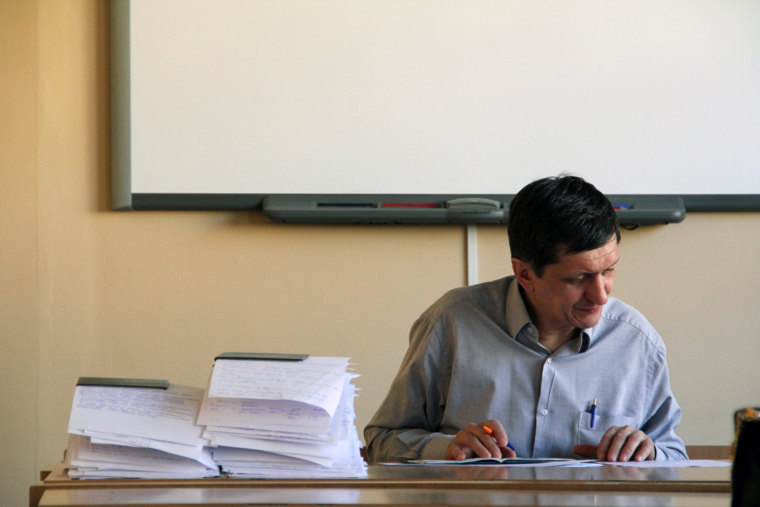 Professor em sua mesa, com quadro ao fundo. Imagem ilustrativa texto Pedagogia.