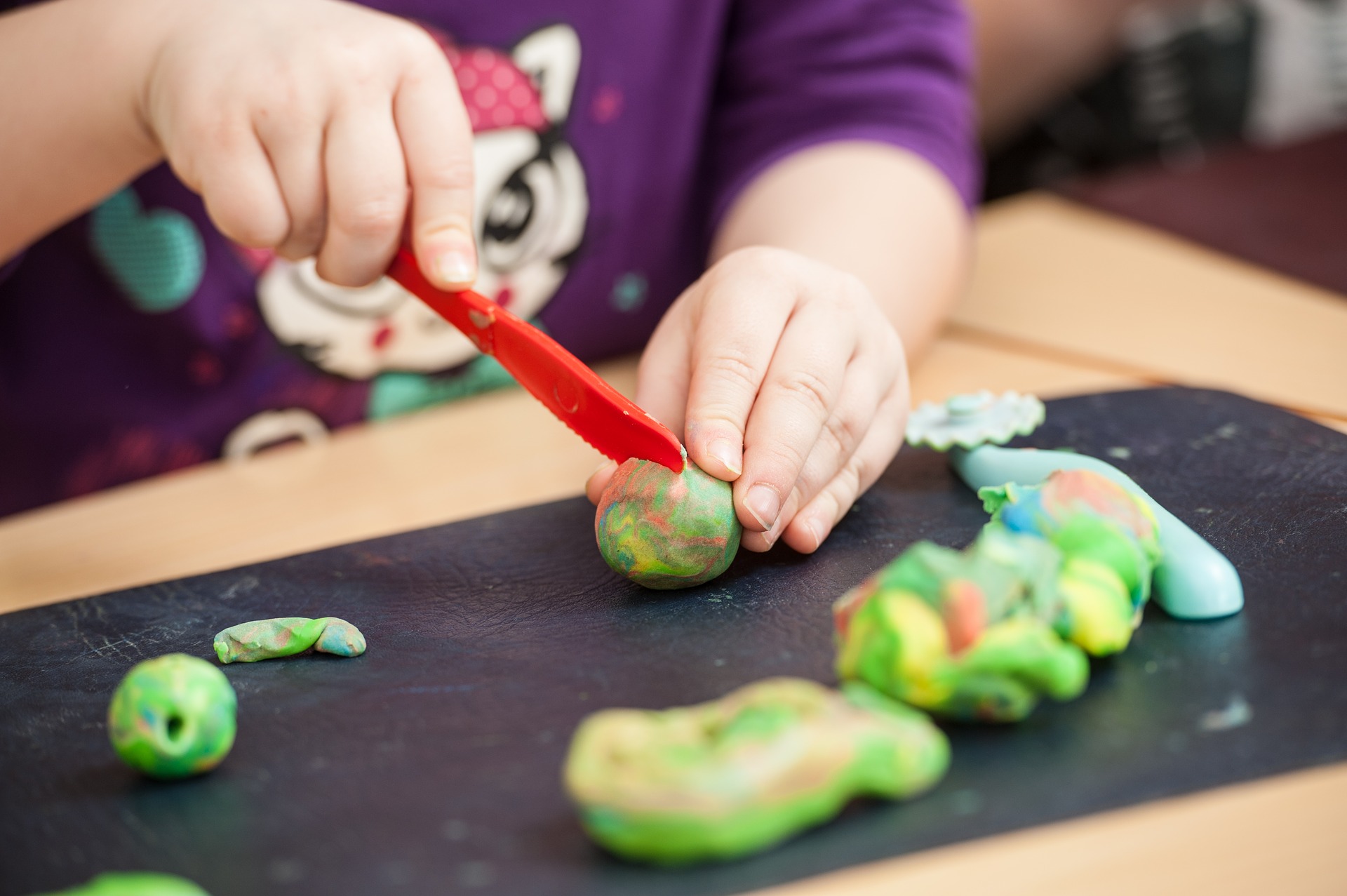 Mãos de criança brincando com faca de plástico e massa de modelar. Imagem ilustrativa texto como fazer massinha.