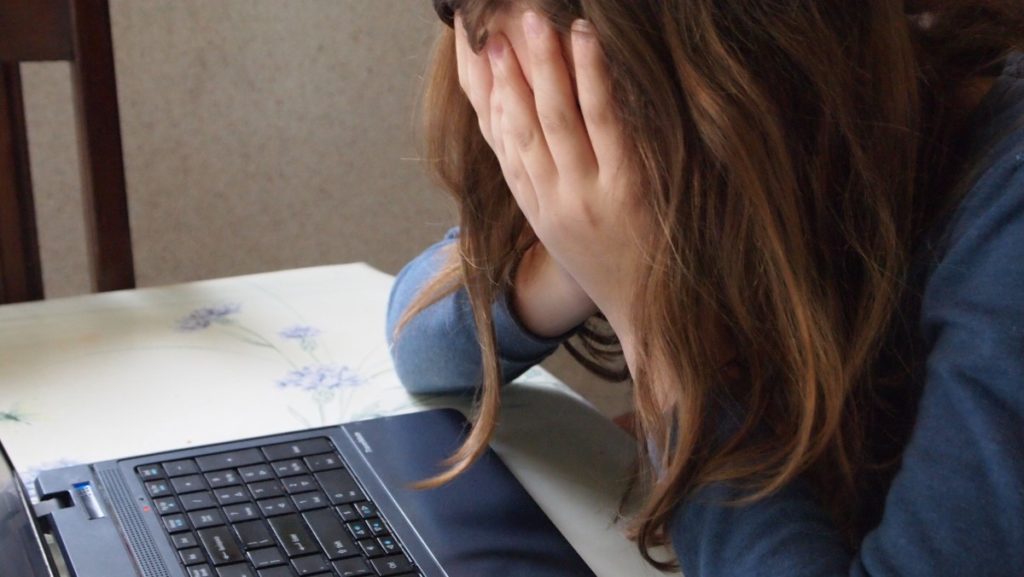 Menina com as mãos no rosto, diante de notebook. Imagem ilustrativa texto saúde mental infantil.