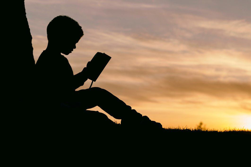 Silhueta de um menino lendo ao pôr do sol. Imagem ilustrativa texto leitor.