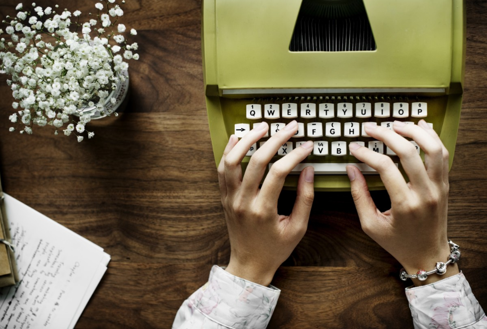 Mão de mulher em máquina de escrever, com flores. Imagem ilustrativa texto escritoras mulheres.