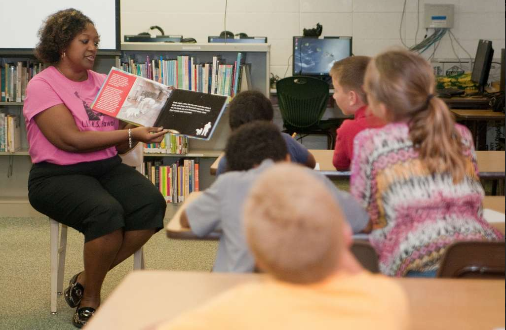 Professora mostrando livro a alunos. Imagem ilustrativa texto como escolher livros para escola.